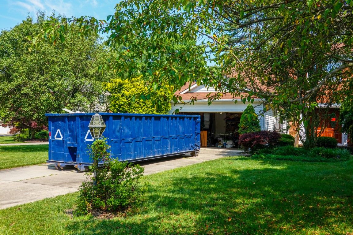 Blue roll-off dumpster parked in a residential driveway beside a lawn and mature trees
