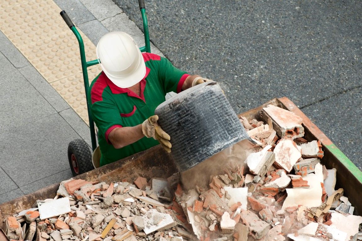Worker in a hard hat moving a bucket of broken brick and demolition rubble with a hand truck