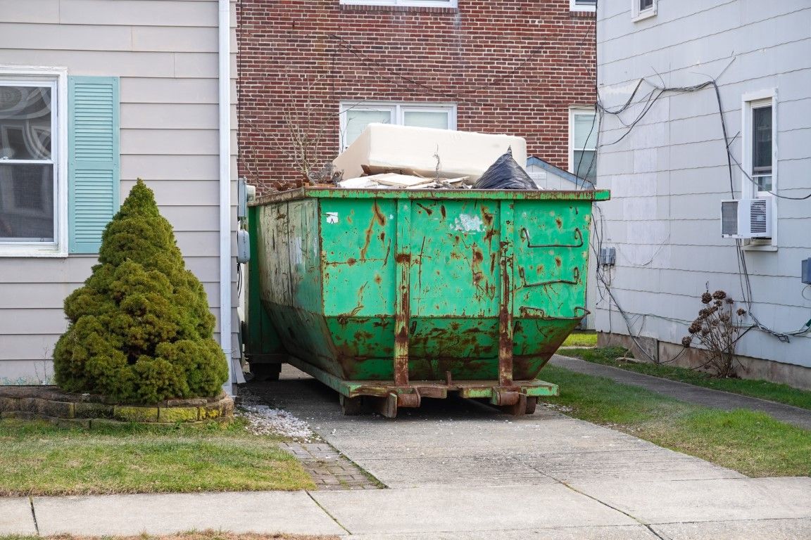 Green roll-off container placed on a paved area beside a house with a shrub in front