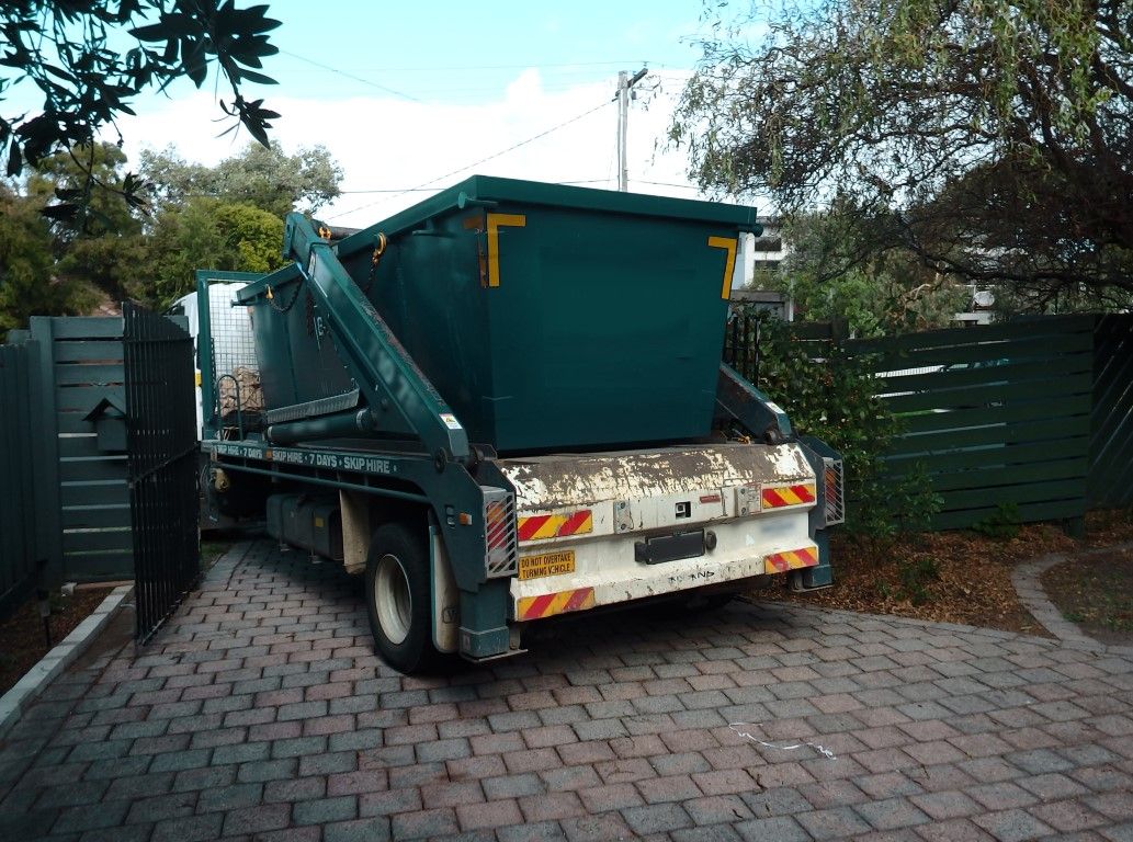 Hook-lift truck backing an empty green roll-off container into a paved yard