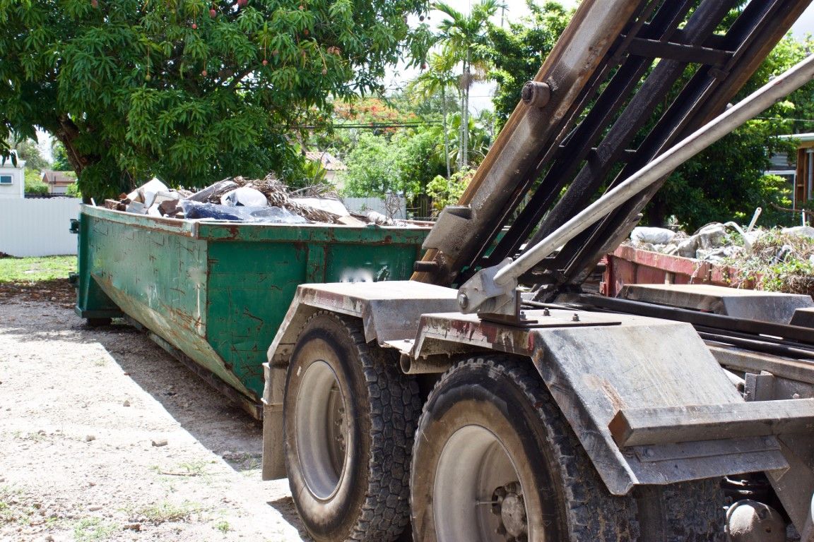 Hook-lift truck carrying a green roll-off container full of debris down a residential street