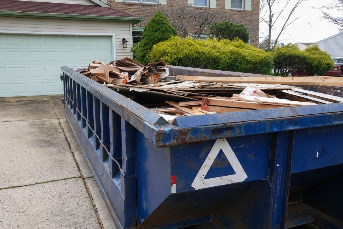 Blue roll-off dumpster loaded with lumber and demolition debris beside a house