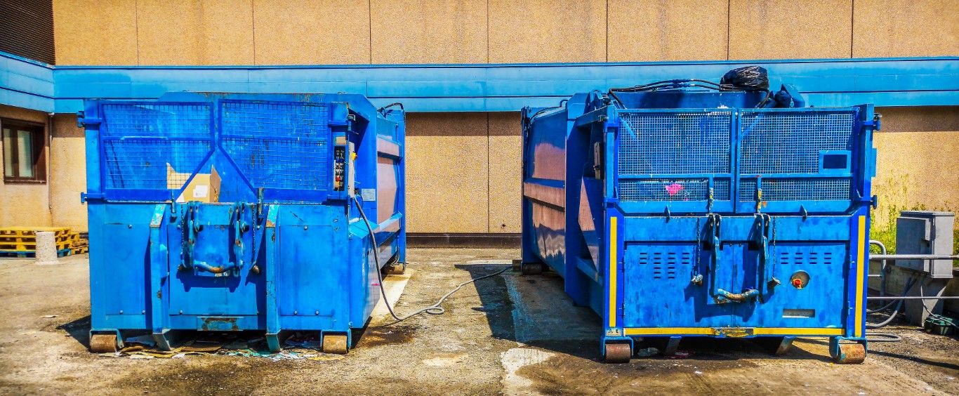 Two large blue dumpster compactor units parked side by side outside a commercial building