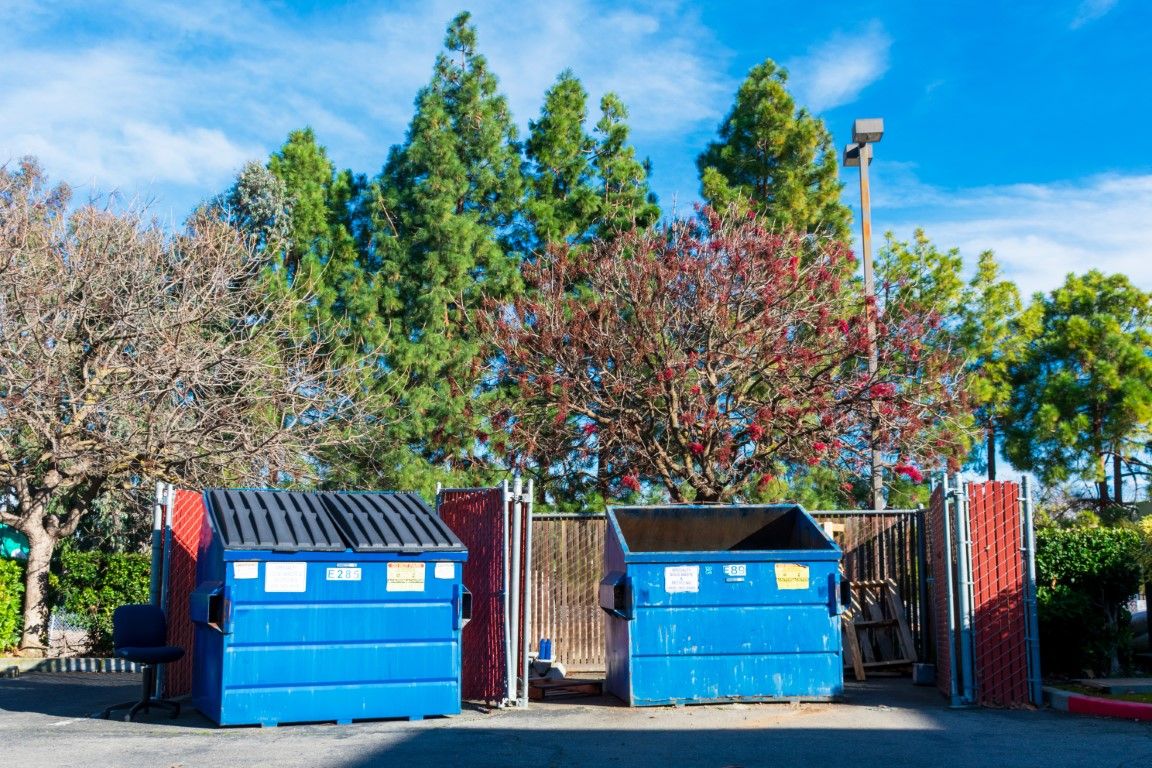 Blue dumpsters behind a red metal fence with trees in the background