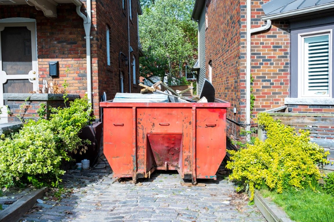 Red dumpster placed on a paved side yard between two brick homes with shrubs