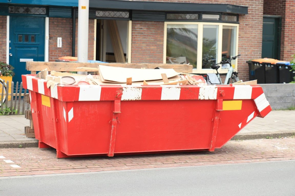 Red and white striped open-top dumpster with wood debris parked on a street in front of a building